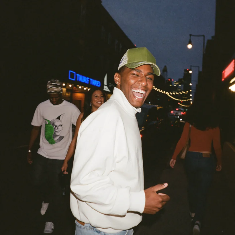 Craig in a white sweater and cap, laughing on a street at night