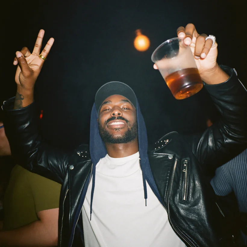 Johnny in a cap and leather jacket, throwing a peace sign and raising a drink